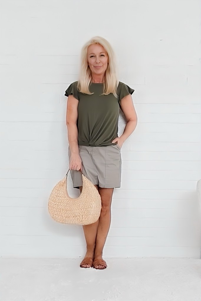 A woman stands against a white wall, smiling while wearing a green top and beige shorts. She holds a straw bag in her right hand.