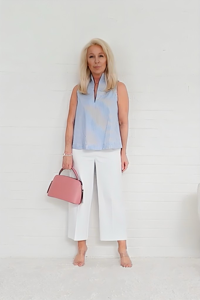 A woman standing in a minimalist setting, wearing a light blue sleeveless top and white wide-legged pants, holding a pink handbag.