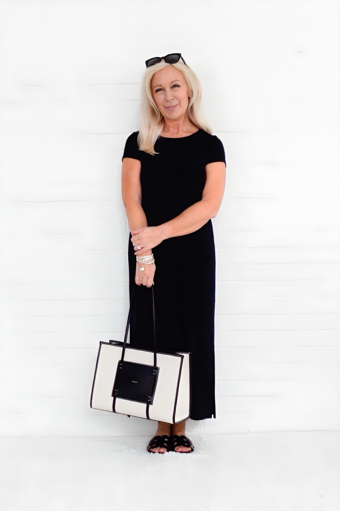 A woman with long blonde hair wearing a black dress, holding a large beige and black handbag, standing against a white backdrop.