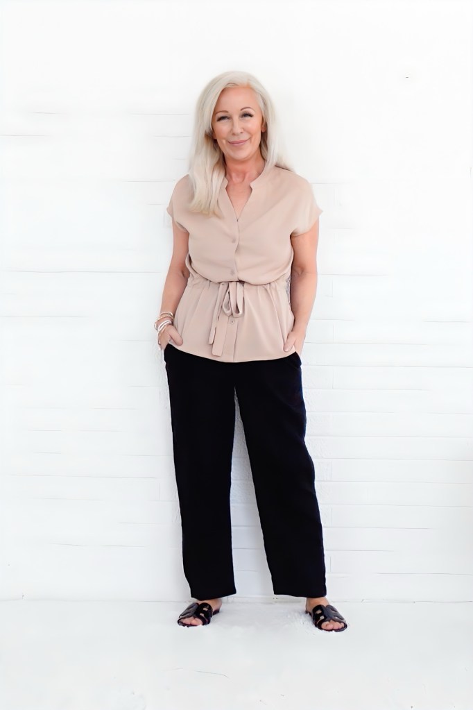 A woman with long, light-colored hair wearing a beige blouse and black pants, standing against a white wooden wall.