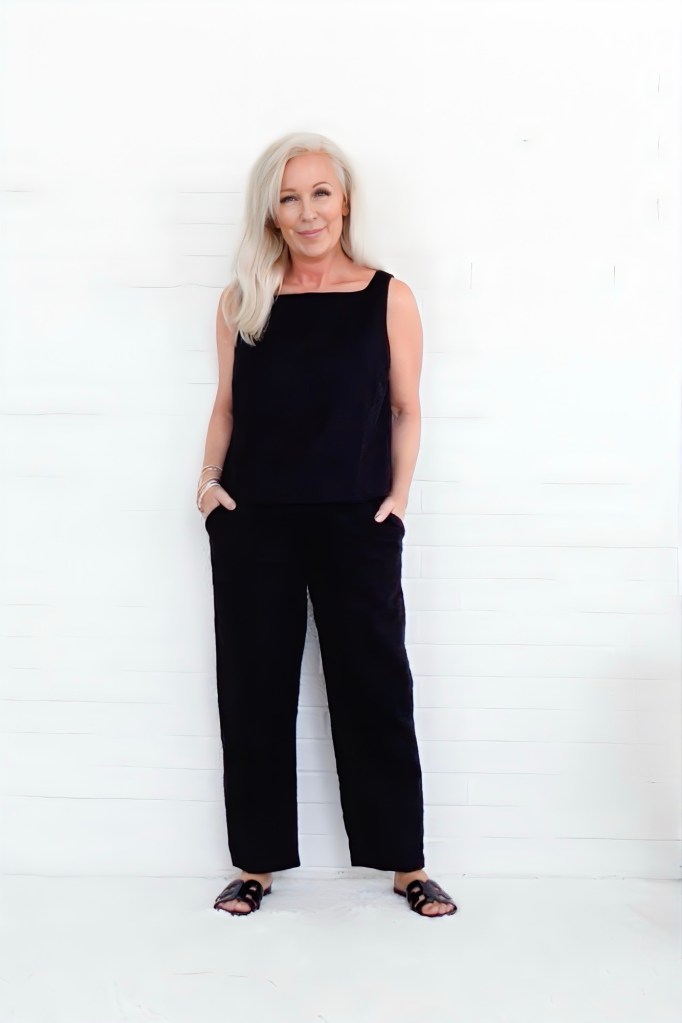 A woman with long blonde hair wearing a black sleeveless top and black pants, standing confidently against a white background.