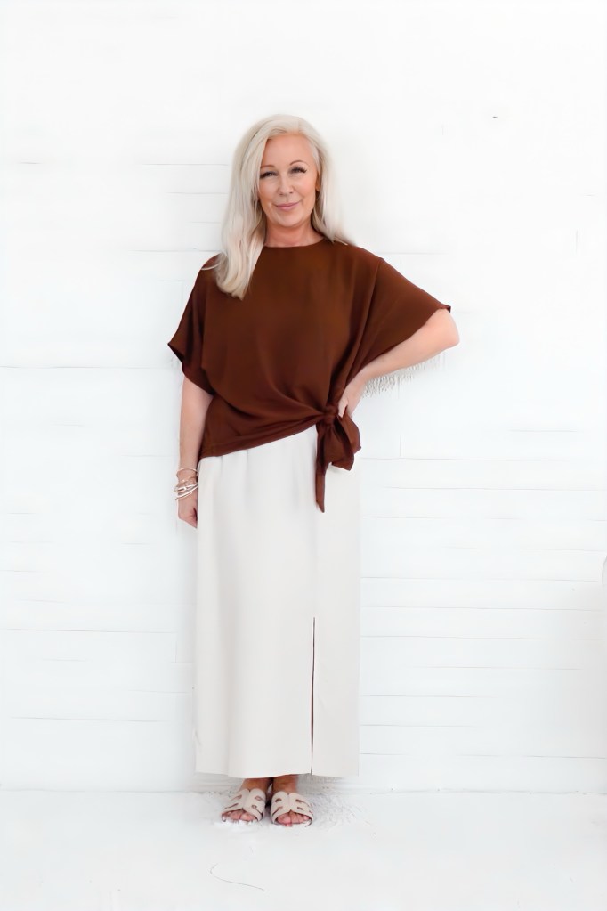 A woman with long blonde hair stands confidently against a white background, wearing a brown top tied at the waist and a beige maxi skirt. She is accessorized with bracelets and wearing light-colored sandals.