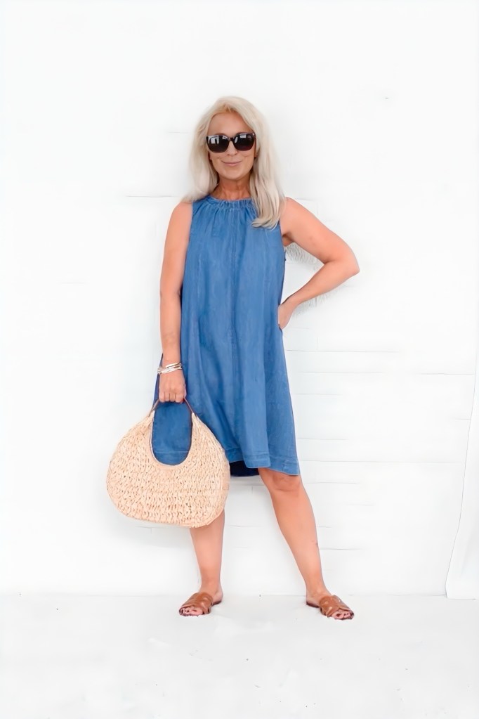 A stylish woman with long blonde hair wearing sunglasses and a denim dress, holding a woven bag, standing against a white background.