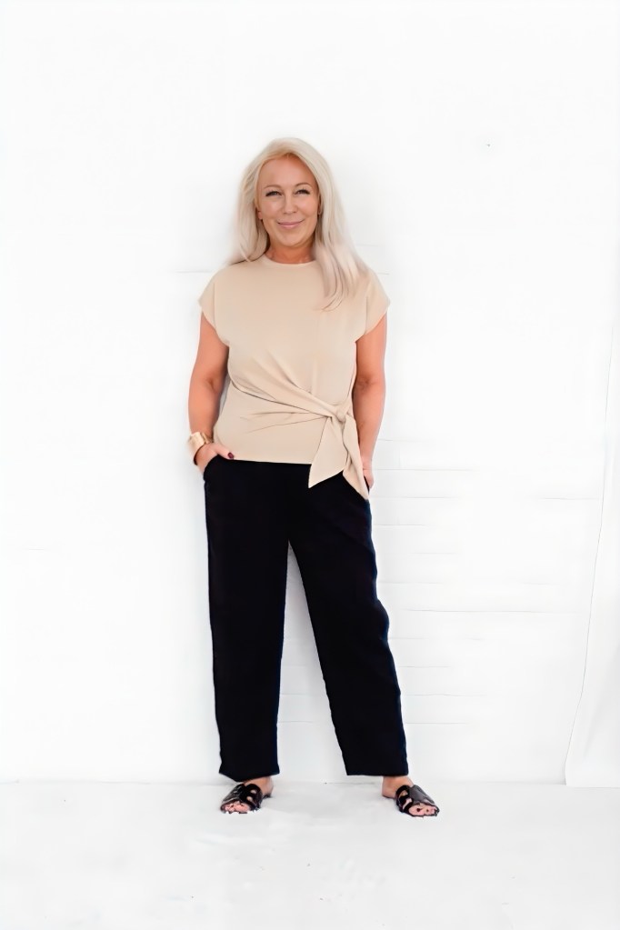 A smiling woman with long blonde hair stands against a white background, wearing a beige top and black pants, posing casually with one hand in her pocket.