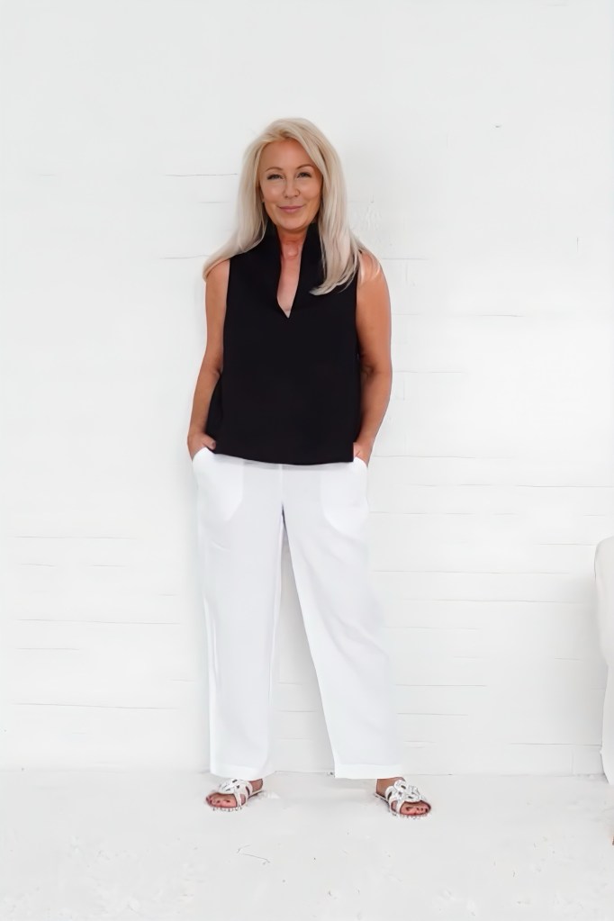 A woman with blonde hair, wearing a black sleeveless top and white wide-leg pants, stands against a white background, smiling and posing with her hands in her pockets.
