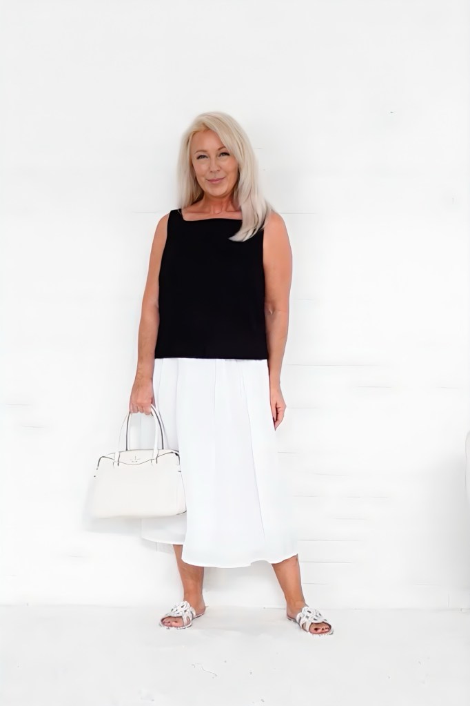 A woman stands smiling in front of a white background, wearing a black sleeveless top, a flowy white skirt, and white sandals. She is holding a light-colored handbag.