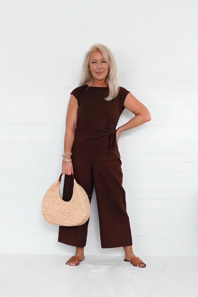 A stylish woman with long blonde hair wearing a brown outfit, holding a straw handbag, stands against a white background.