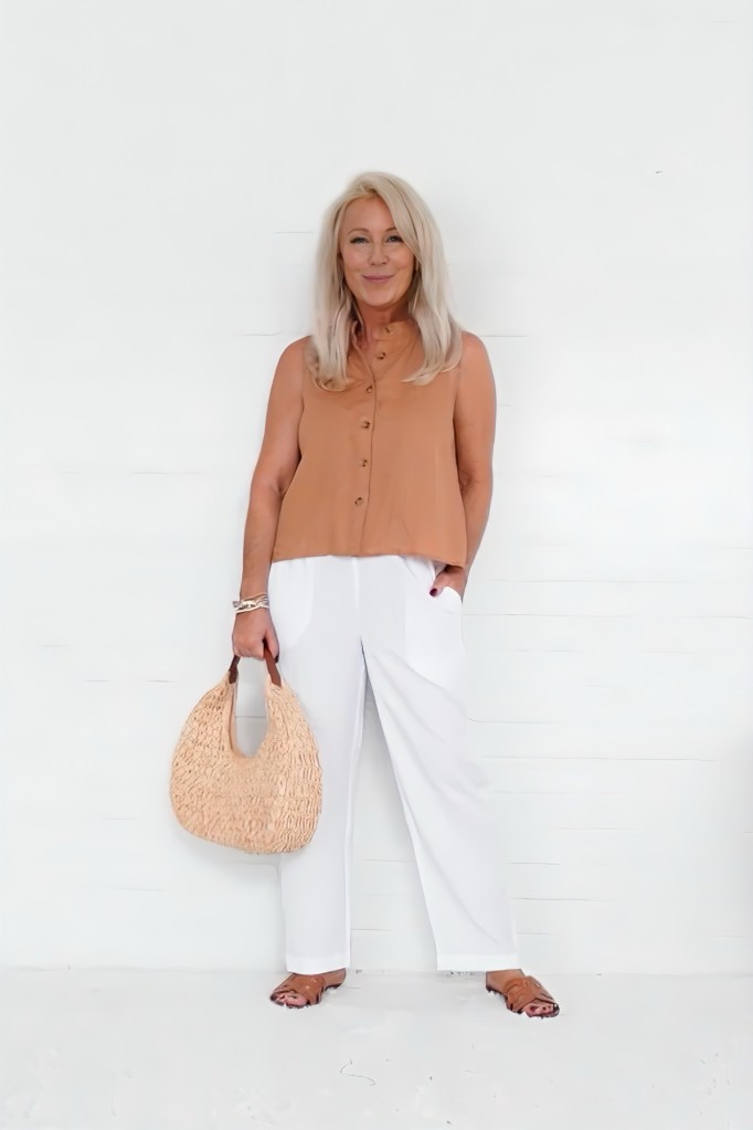 A woman standing in front of a white background wearing a tan sleeveless top and white pants, holding a straw bag.