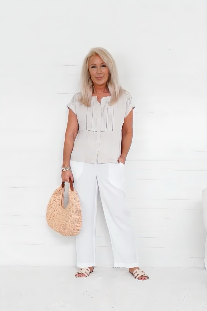 A woman standing in front of a white wall, wearing a light-colored blouse and white pants, holding a woven bag.