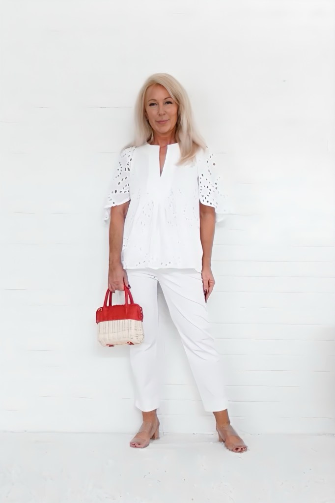 A woman with long blonde hair is standing against a white background, wearing a white top with eyelet details and white pants. She holds a small red and beige handbag.