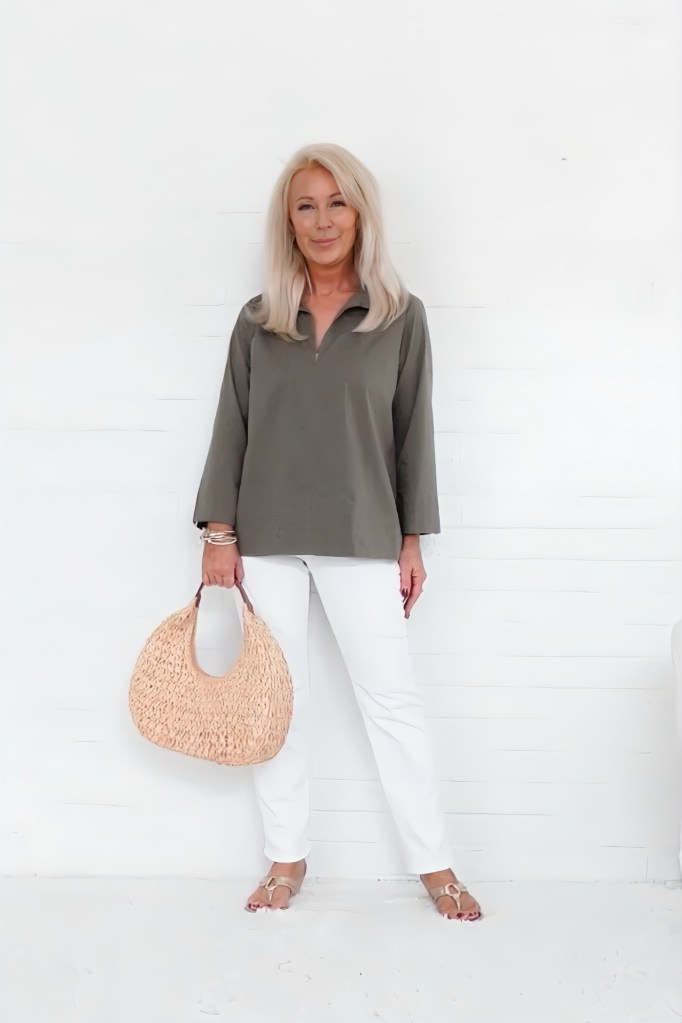 A woman standing against a white wall, wearing a loose olive green top and white pants, holding a woven beige handbag.