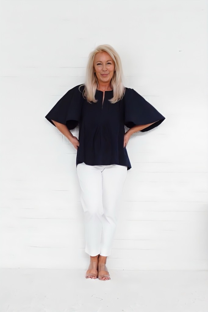 A woman with long blonde hair is standing against a white background, wearing a navy top with wide sleeves and white pants.
