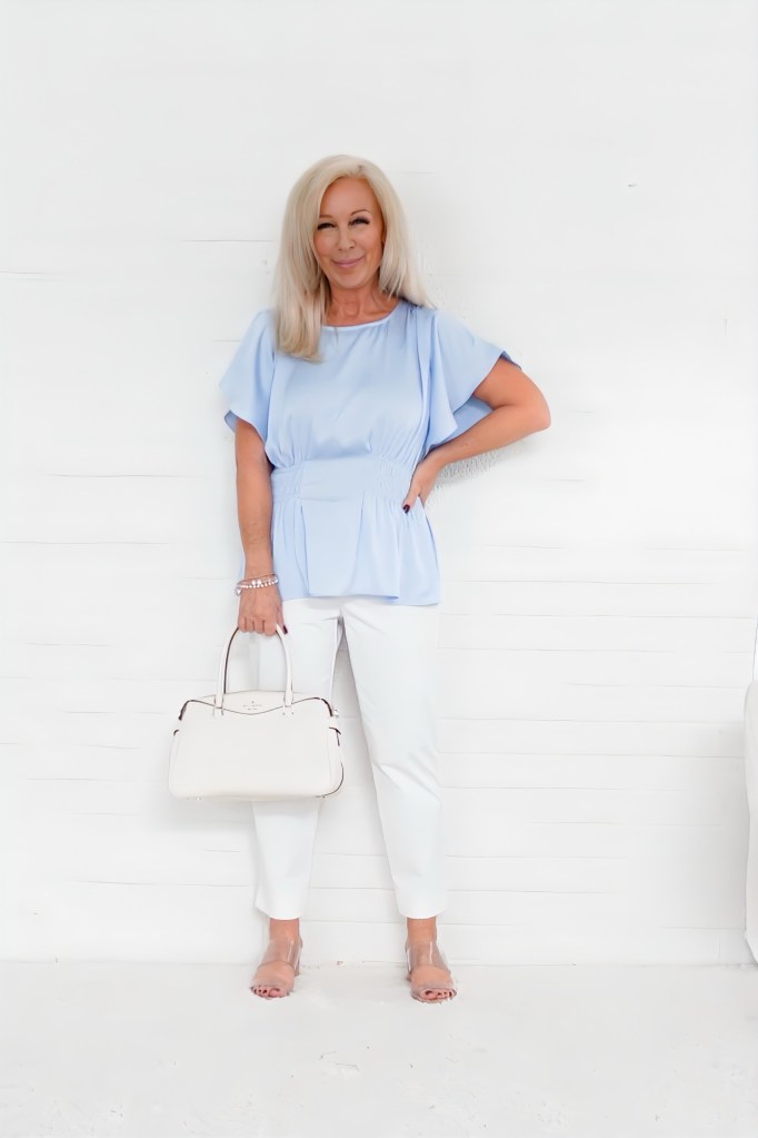 A woman with long blonde hair wearing a light blue blouse and white pants, holding a white handbag, posed in front of a white backdrop.