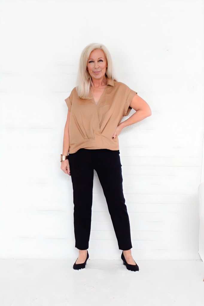 A woman wearing a beige top and black pants, standing against a white background, smiling confidently.