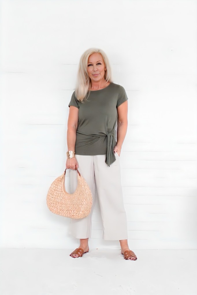 A woman standing against a white background, wearing an olive green top and cream-colored wide-leg pants, holding a woven handbag.