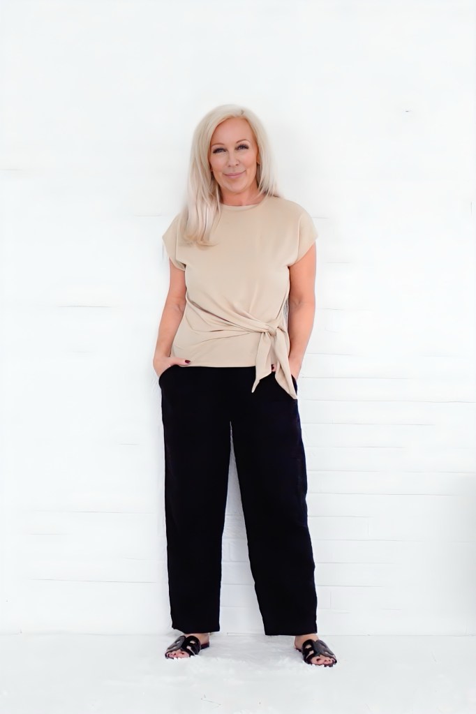 A woman with long blonde hair stands against a white background, wearing a beige top tied at the waist and black wide-leg pants.