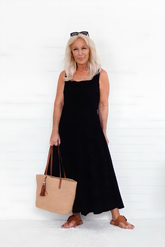 A woman with long blonde hair wearing a black sleeveless dress, holding a tan tote bag and standing in front of a white wall.