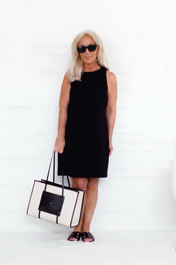 A woman with long blonde hair wearing a black dress and sunglasses, holding a large tote bag, standing in front of a white background.