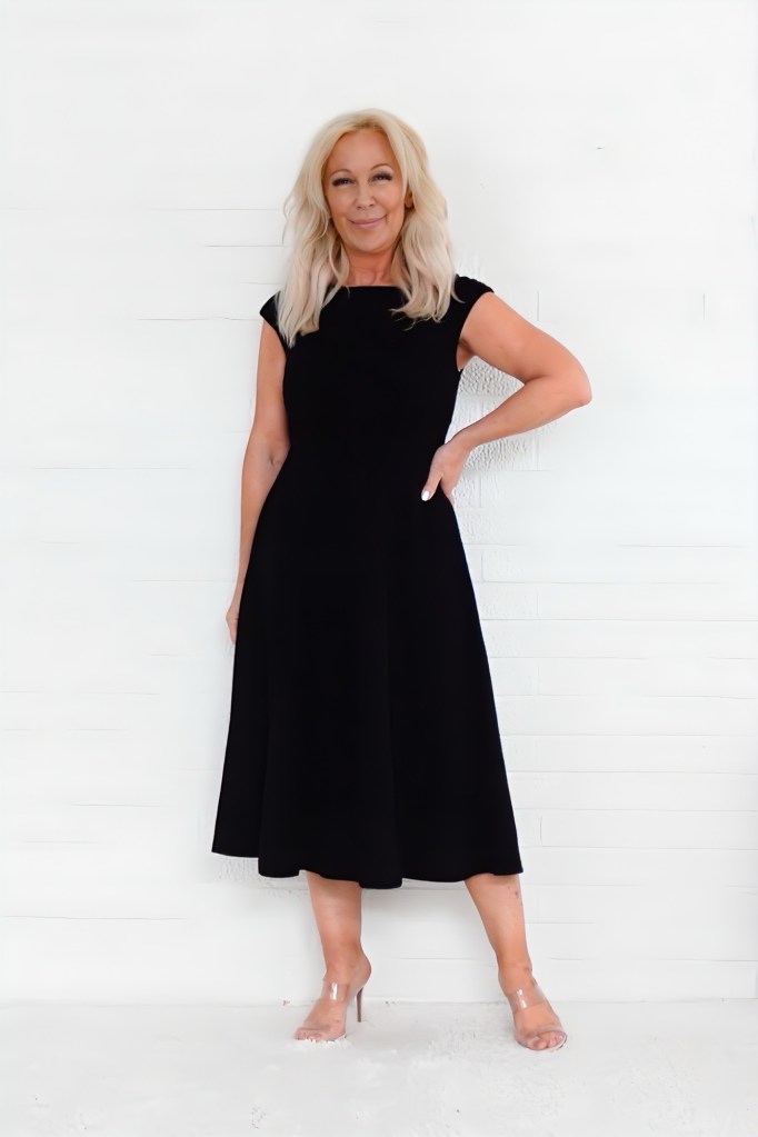 A woman wearing a black dress poses confidently with one hand on her hip against a white background.