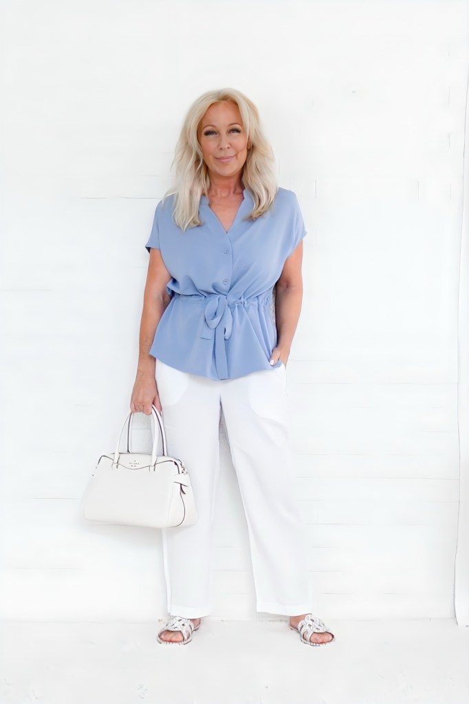 A woman with long blonde hair wearing a blue blouse and white pants, holding a white handbag, standing against a plain white background.