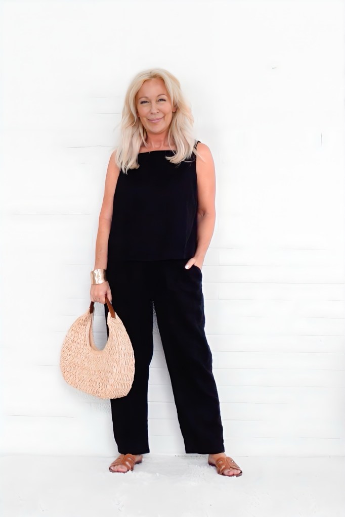 A woman with long blonde hair wearing a black outfit, holding a straw handbag, standing against a white wall.