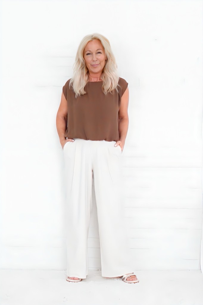 A woman with long blonde hair wearing a brown top and white wide-leg pants, standing against a white background.