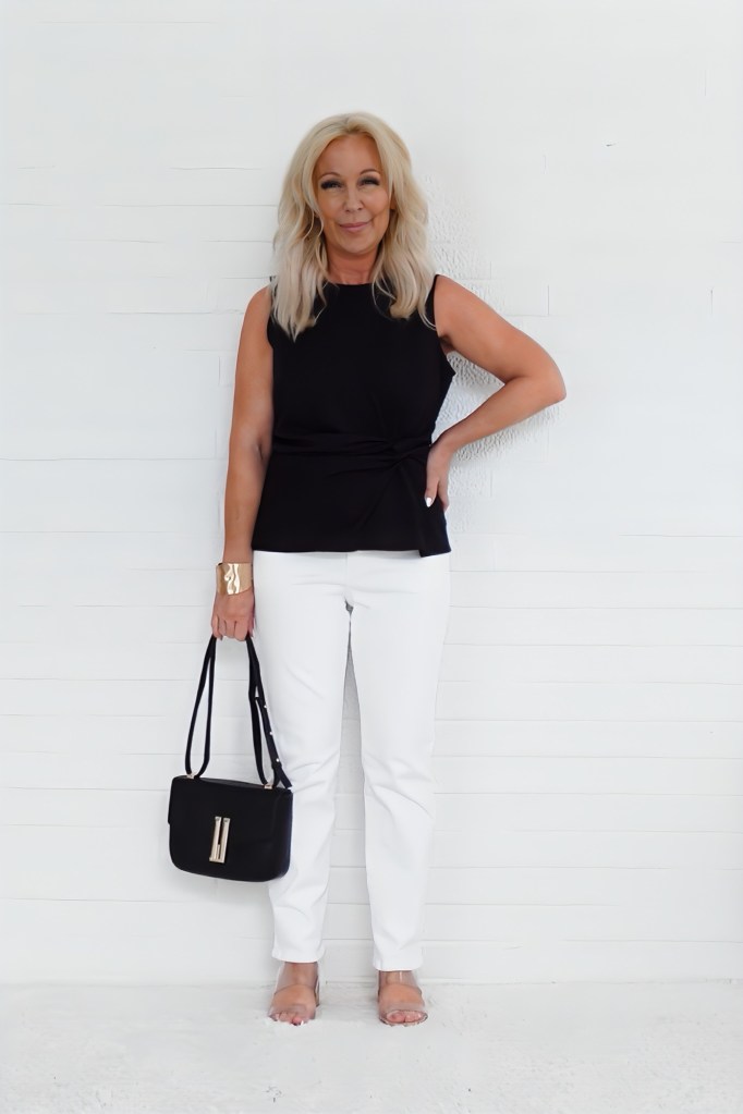 A woman with blonde hair stands smiling against a white wall, wearing a black sleeveless top and white pants, holding a black handbag.