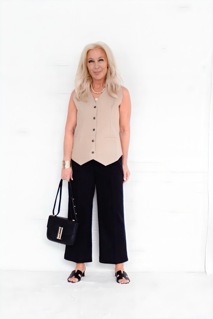 A woman with long blonde hair is standing against a white background, wearing a sleeveless beige top, black wide-leg pants, and black sandals. She holds a black handbag and is accessorized with a pearl necklace.