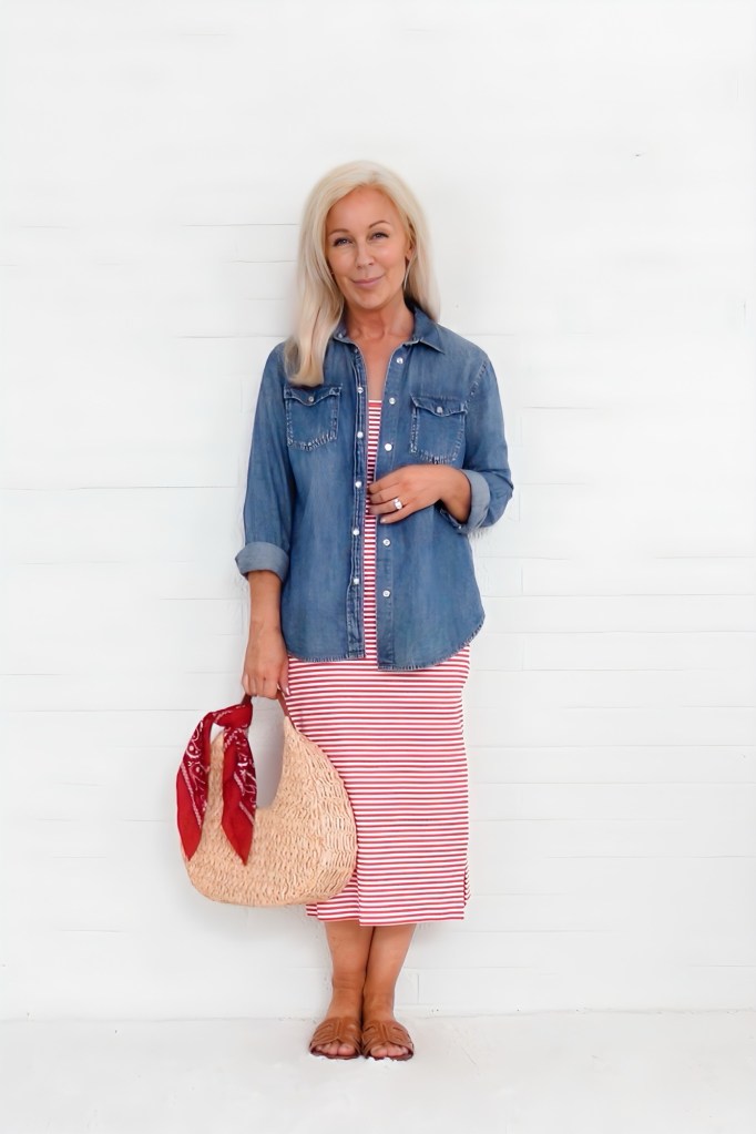 Woman standing against a white background, wearing a denim shirt over a red and white striped dress, holding a straw bag with a red patterned scarf.