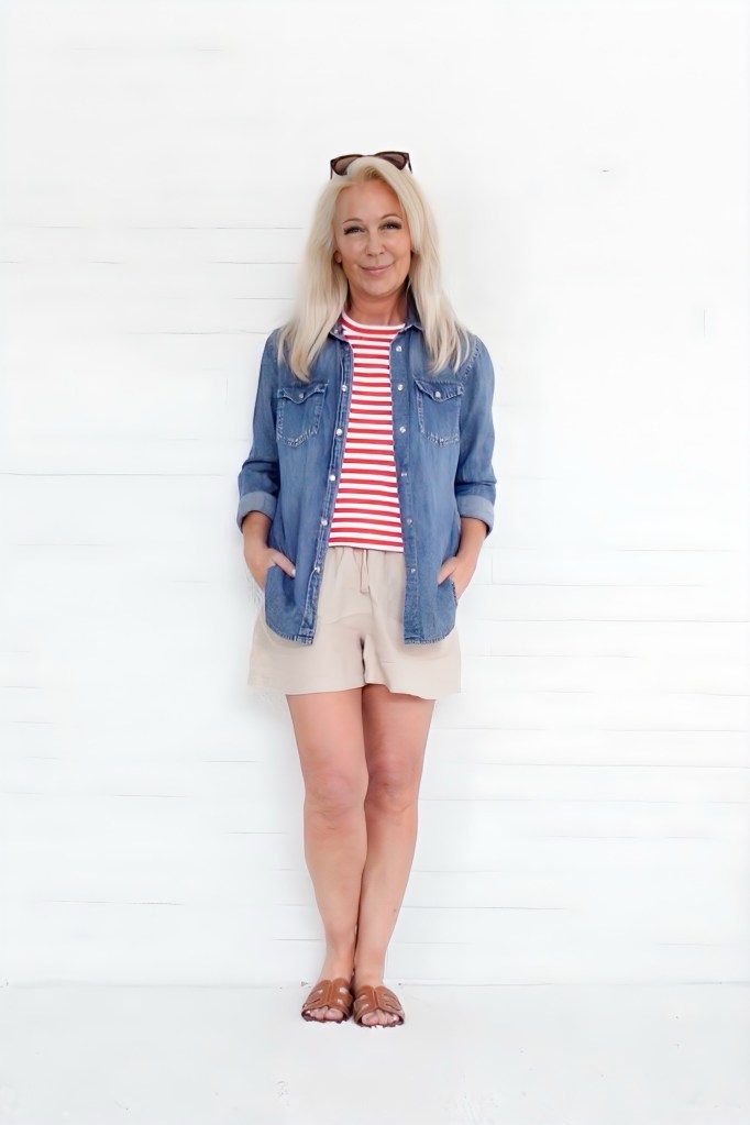 A woman with long blonde hair stands against a white wall, wearing a denim jacket over a red and white striped shirt and beige shorts, with sandals on her feet.
