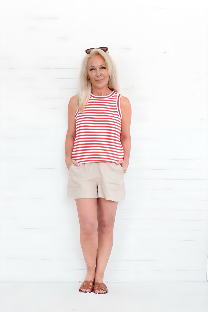 A woman with long blonde hair is standing against a white wall, wearing a red and white striped tank top and beige shorts. She has a relaxed smile and is holding her hands in the pockets of her shorts.