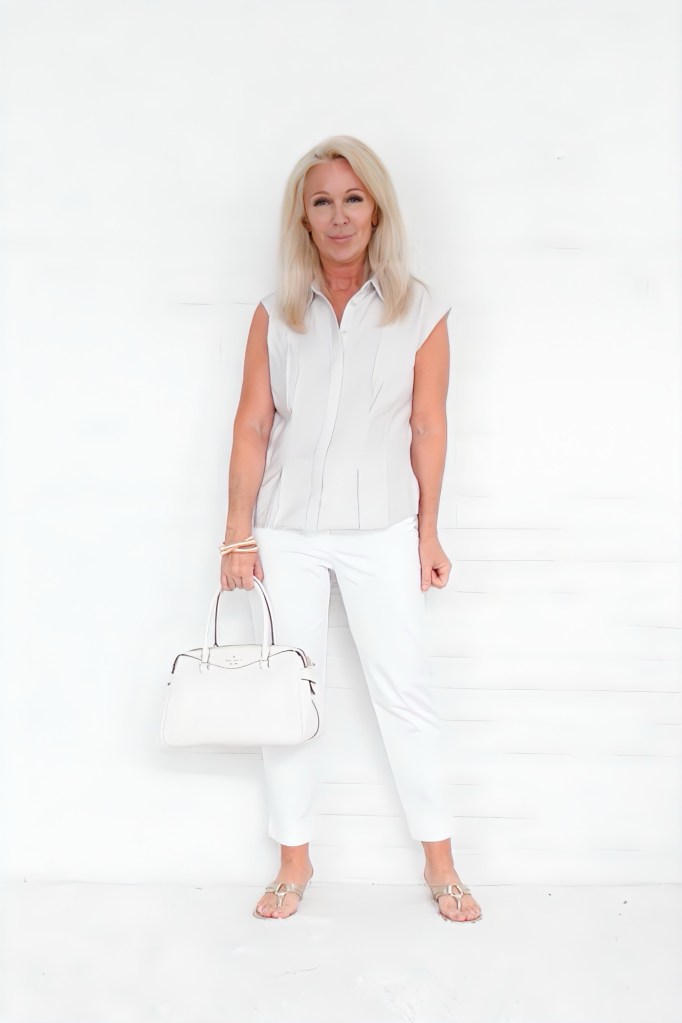 A woman standing against a white background, wearing a light gray sleeveless blouse, white pants, and holding a white handbag. She has long, blonde hair and is smiling.