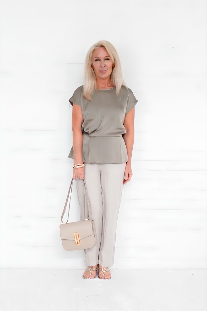 A woman standing against a white background, wearing a stylish olive green top and light-colored trousers, holding a beige handbag and looking confidently at the camera.
