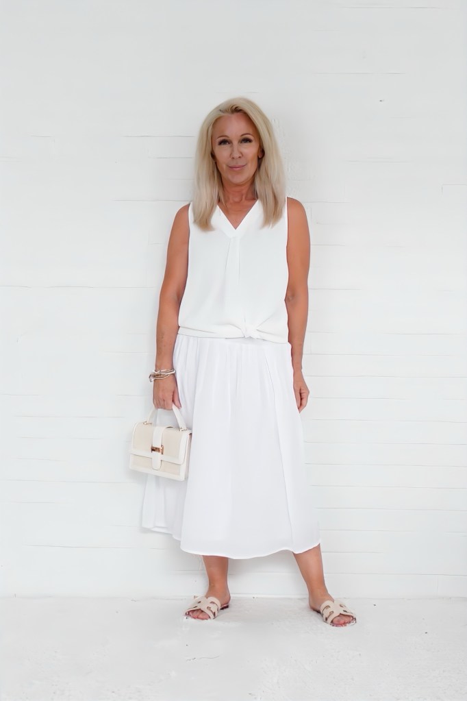 A woman standing against a white wall, wearing a white sleeveless top and a white skirt, holding a small cream handbag. She has long blonde hair and a confident expression.