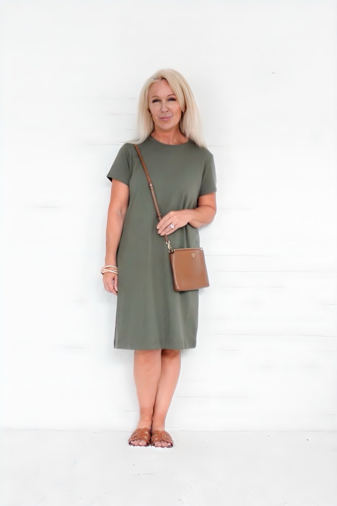 A woman stands against a white wall wearing a lightweight olive green dress, carrying a small brown handbag, with sandals on her feet and a casual smile.