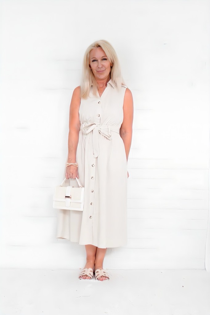 A woman standing against a white backdrop, wearing a light beige sleeveless dress with buttons and a tie belt, holding a stylish white handbag.