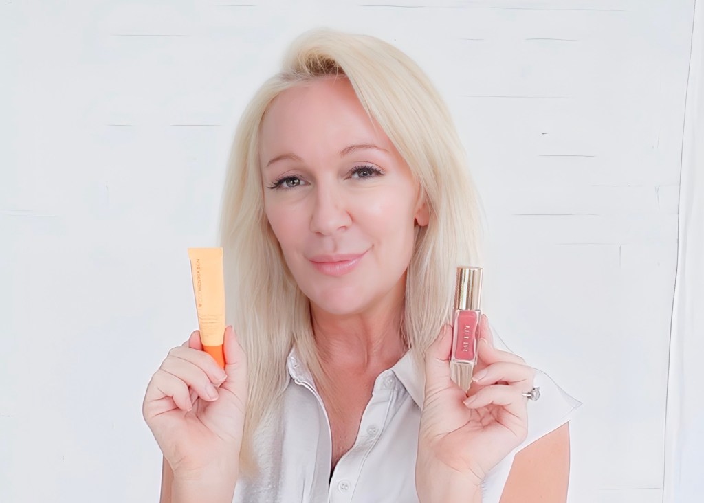 A woman with blonde hair holds a sunscreen product in one hand and a lip gloss in the other, smiling at the camera against a light background.