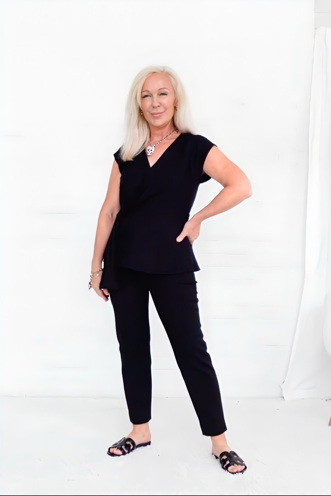 A woman with long blonde hair poses confidently in a black outfit, featuring a fitted top and tailored pants, against a plain white background.