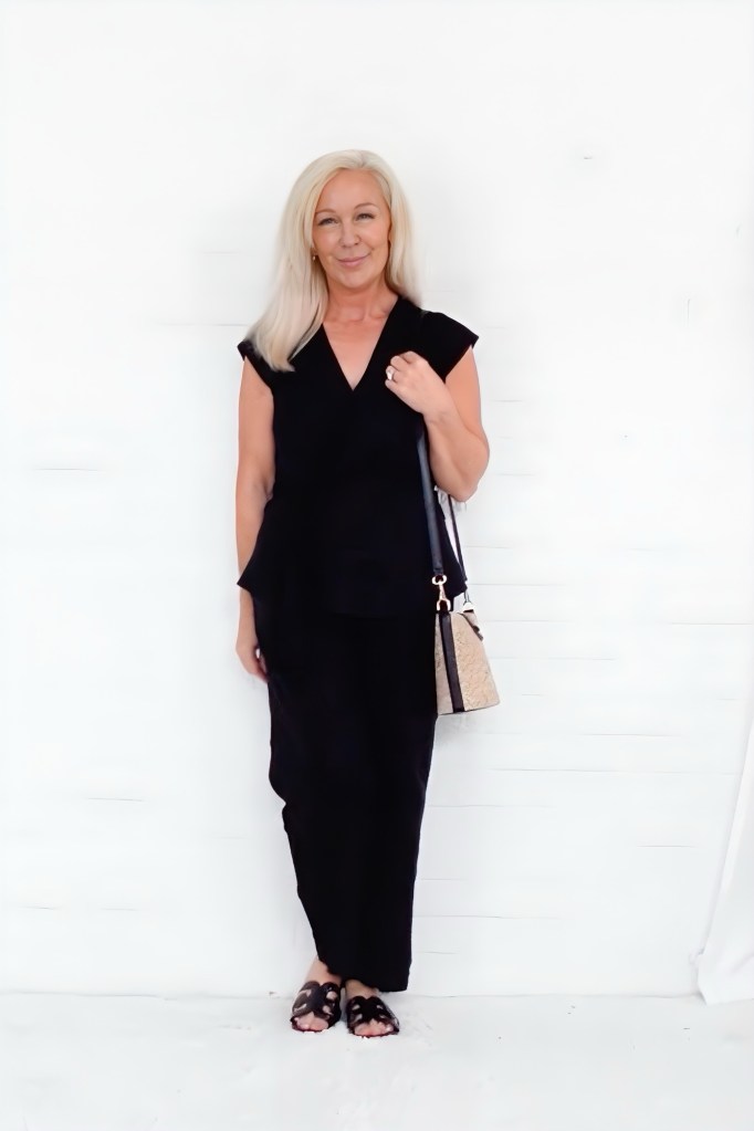 A woman standing against a white background, wearing a black outfit with a stylish cut, holding a small bag. She has long blonde hair and is smiling at the camera.