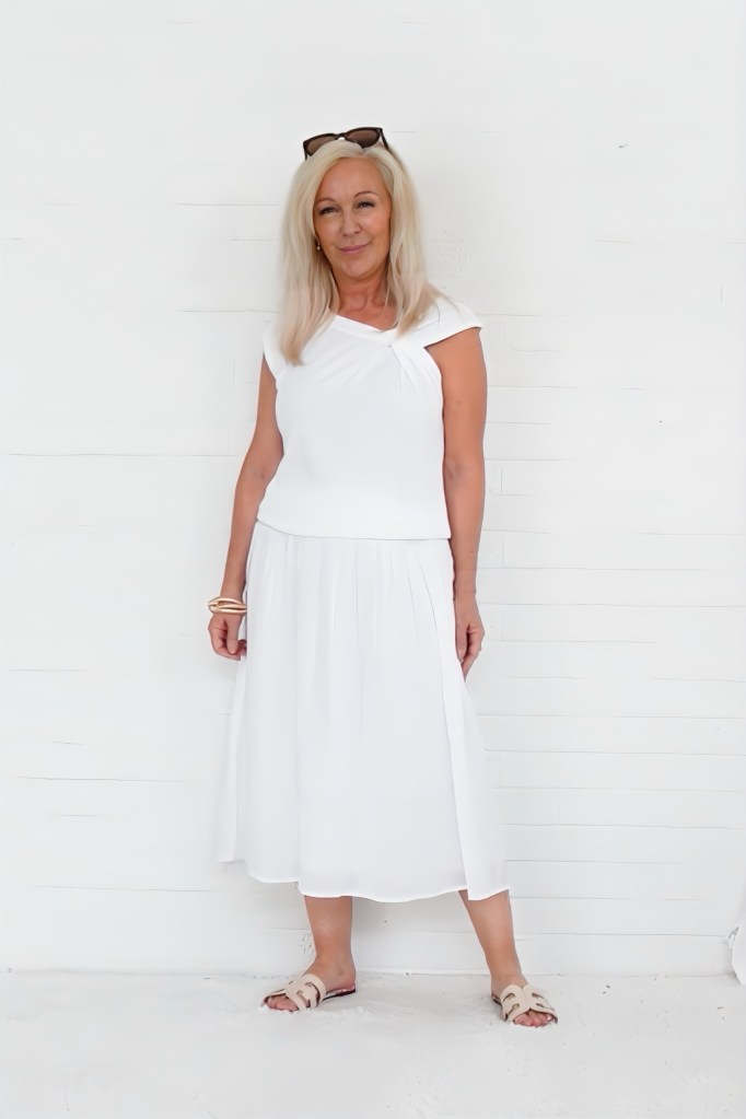 A woman standing against a white wall, wearing a white top and a matching skirt, with blonde hair and sunglasses resting on her head.