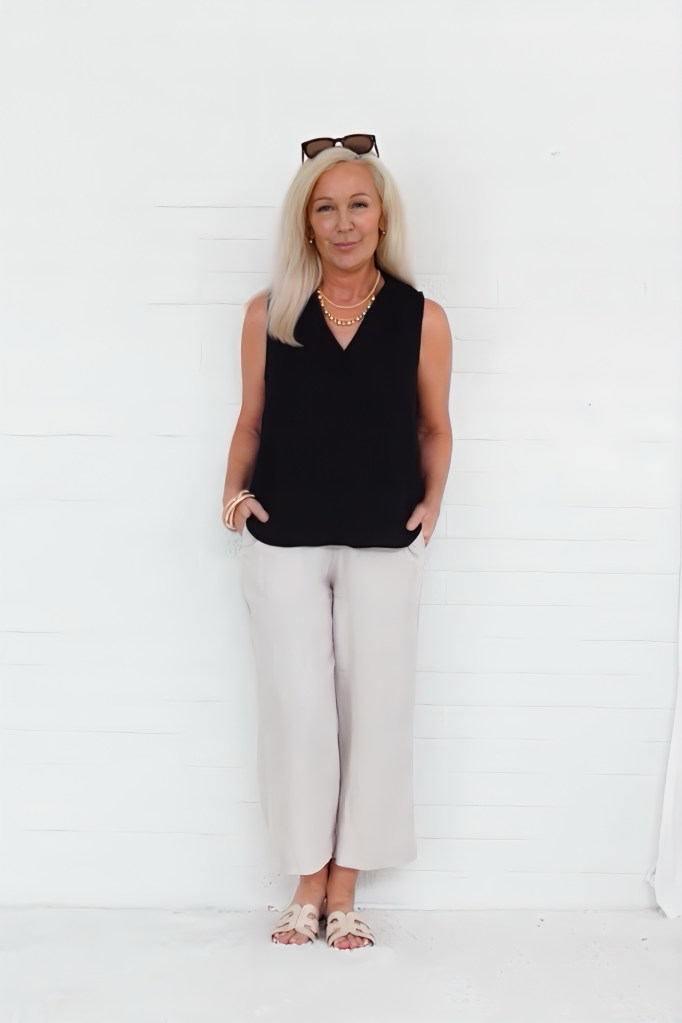 A woman with long blonde hair stands in front of a white wall, wearing a black sleeveless top, light beige wide-leg pants, and sandals, with sunglasses on her head and a necklace.