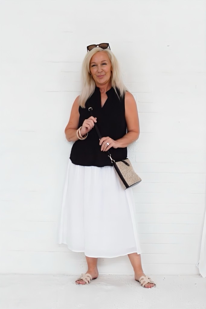 A woman with long blonde hair smiles while standing in front of a white wall, wearing a black sleeveless top, a white flowy skirt, and beige sandals. She holds a small handbag and has sunglasses atop her head.