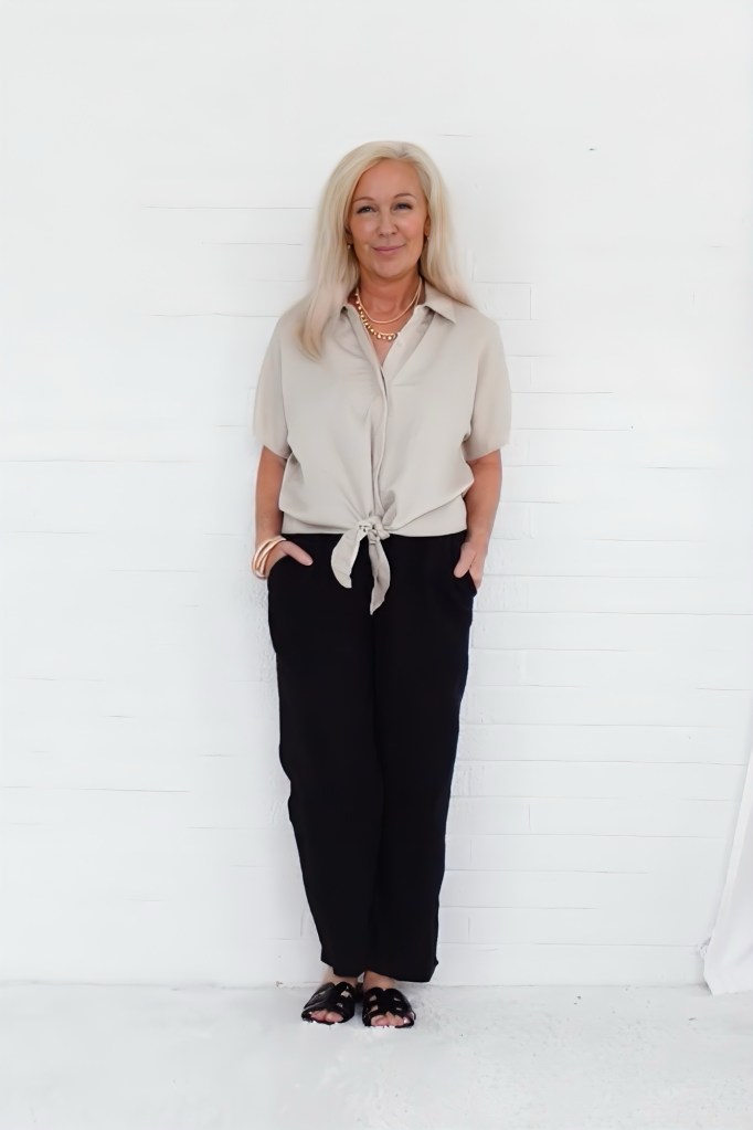 A woman with long blonde hair stands against a white background, wearing a knotted light beige shirt and black pants, accessorized with bracelets and a necklace.