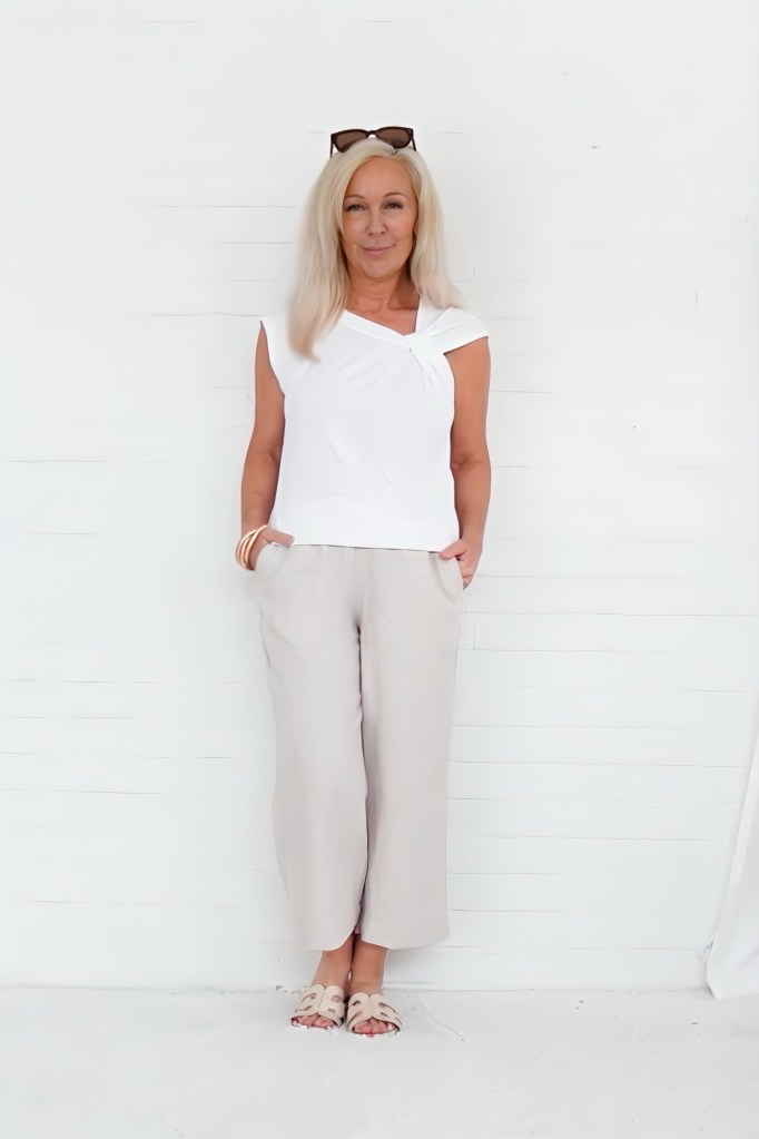 A woman with long blonde hair stands against a white background, wearing a white top and beige wide-leg pants, with sunglasses on her head.