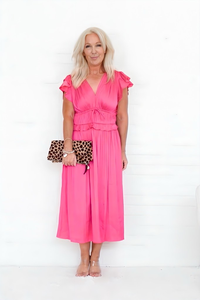 A woman wearing a bright pink dress with ruffled sleeves stands against a white background, holding a leopard print clutch and smiling.