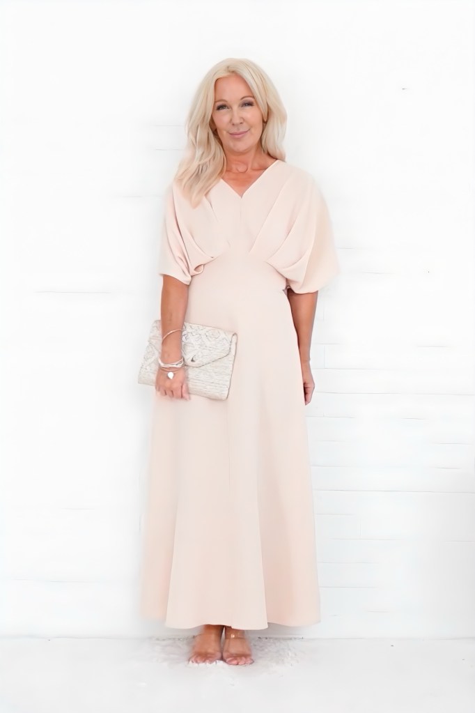 A woman with long blonde hair wearing a pale pink dress and holding a clutch purse, standing against a white backdrop.