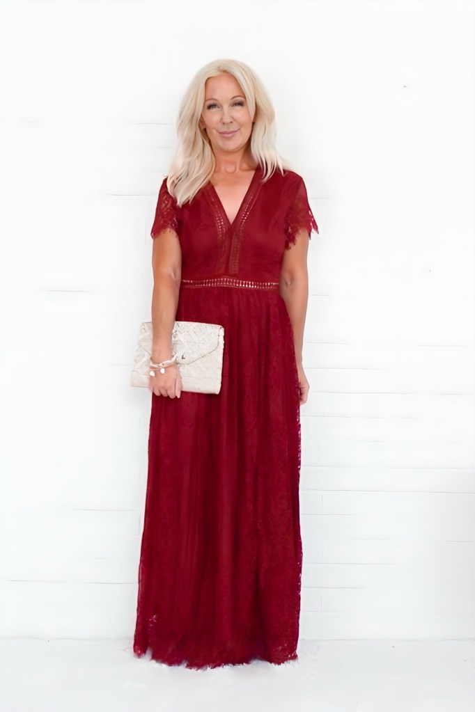 A woman in a red lace maxi dress holding a beige clutch, standing in front of a white background with a friendly smile.