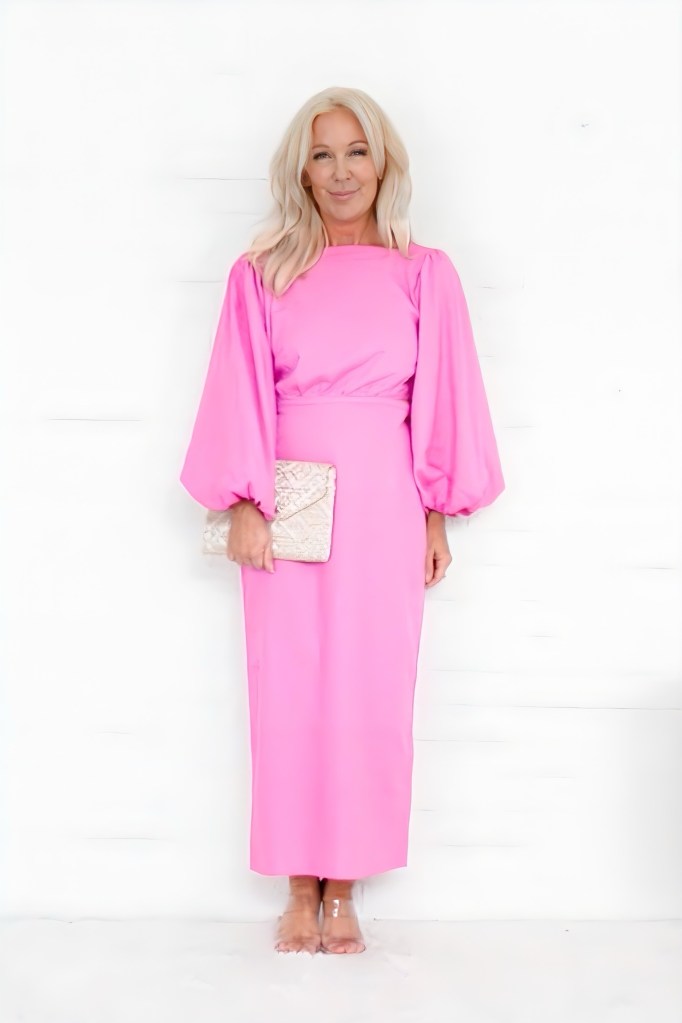 A woman smiling in a long pink dress with puffy sleeves, holding a small beige clutch, standing against a white background.