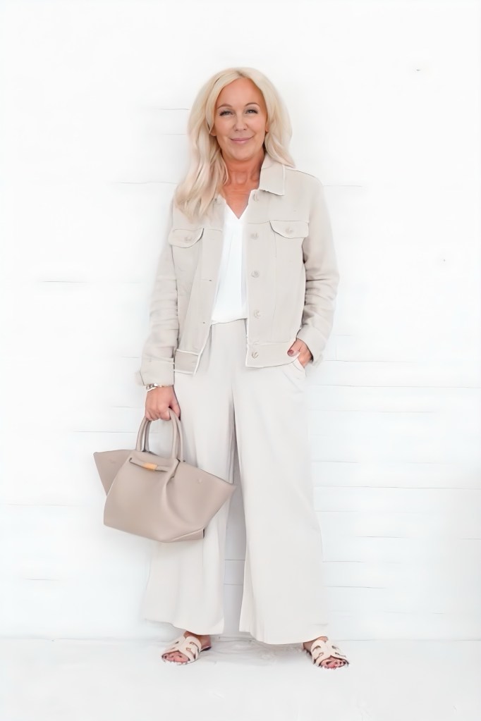 A woman standing against a white background, wearing a light beige jacket and wide-legged beige pants, holding a stylish gray handbag.