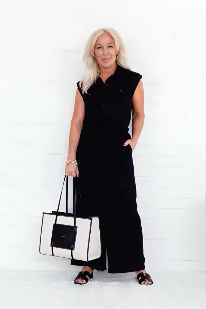 A woman wearing a black jumpsuit with cap sleeves, holding a white handbag with black accents, standing against a white backdrop.